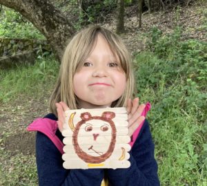 A child holds up an art project with a monkey painted on wood, with Tilden Park in background. Taken at Monkey Business Camp Summer 2022