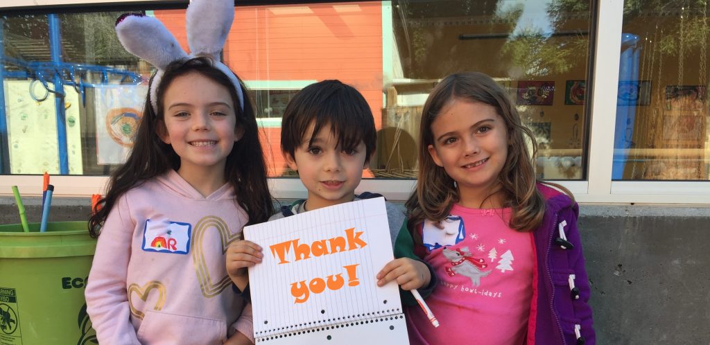 Three children smile, one wearing bunny ears and another holding a notebook that says "thank you!"