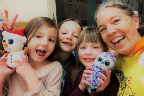 Heather Mitchell, founding director of Monkey Business Camp, smiles with three children, two of whom hold up stuffed toys while the third gives the peace symbol with her hand